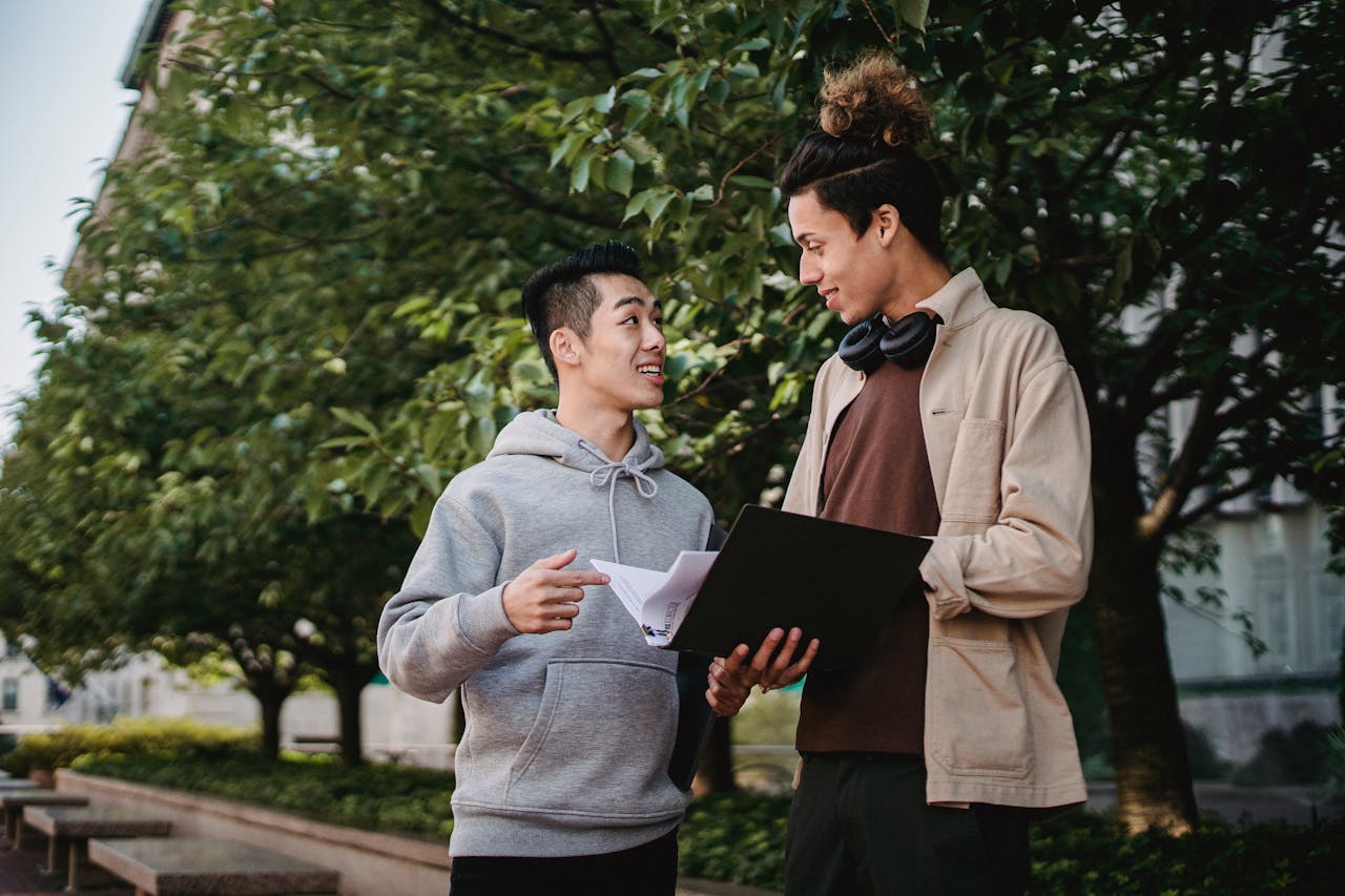 Two diverse young men engaged in a friendly discussion outdoors, surrounded by greenery on campus.