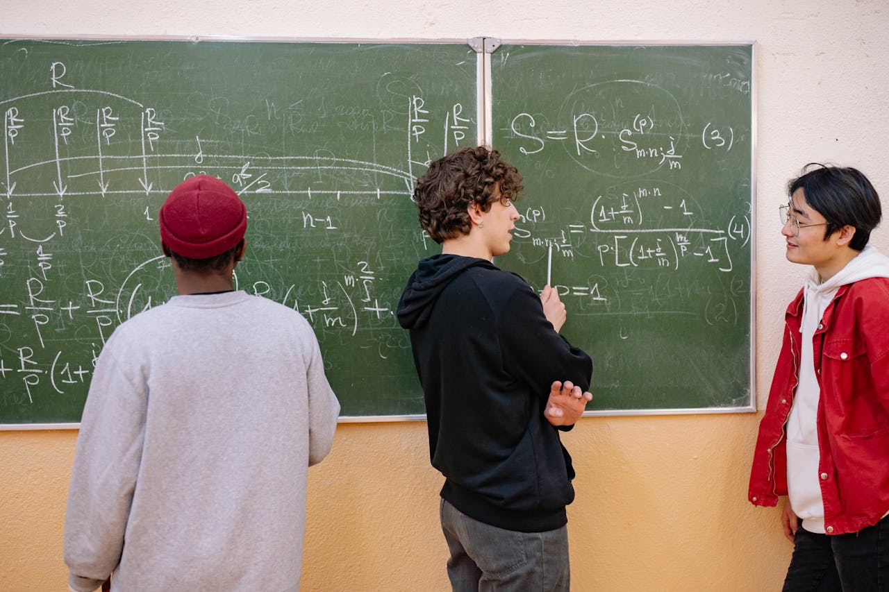 Three students discuss math equations on a classroom blackboard, fostering learning and teamwork.